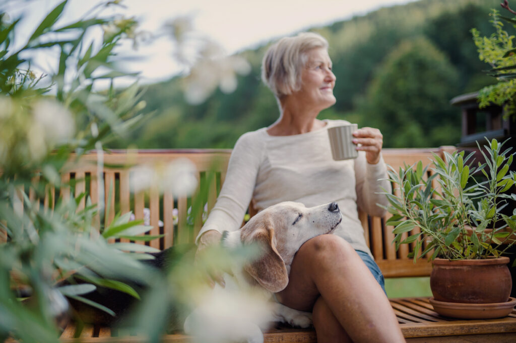 Das Bild zeigt eine ältere Dame mit einer Tasse Tee und ihrem Hund. Sie sitzt draußen auf ihrer Terrasse.